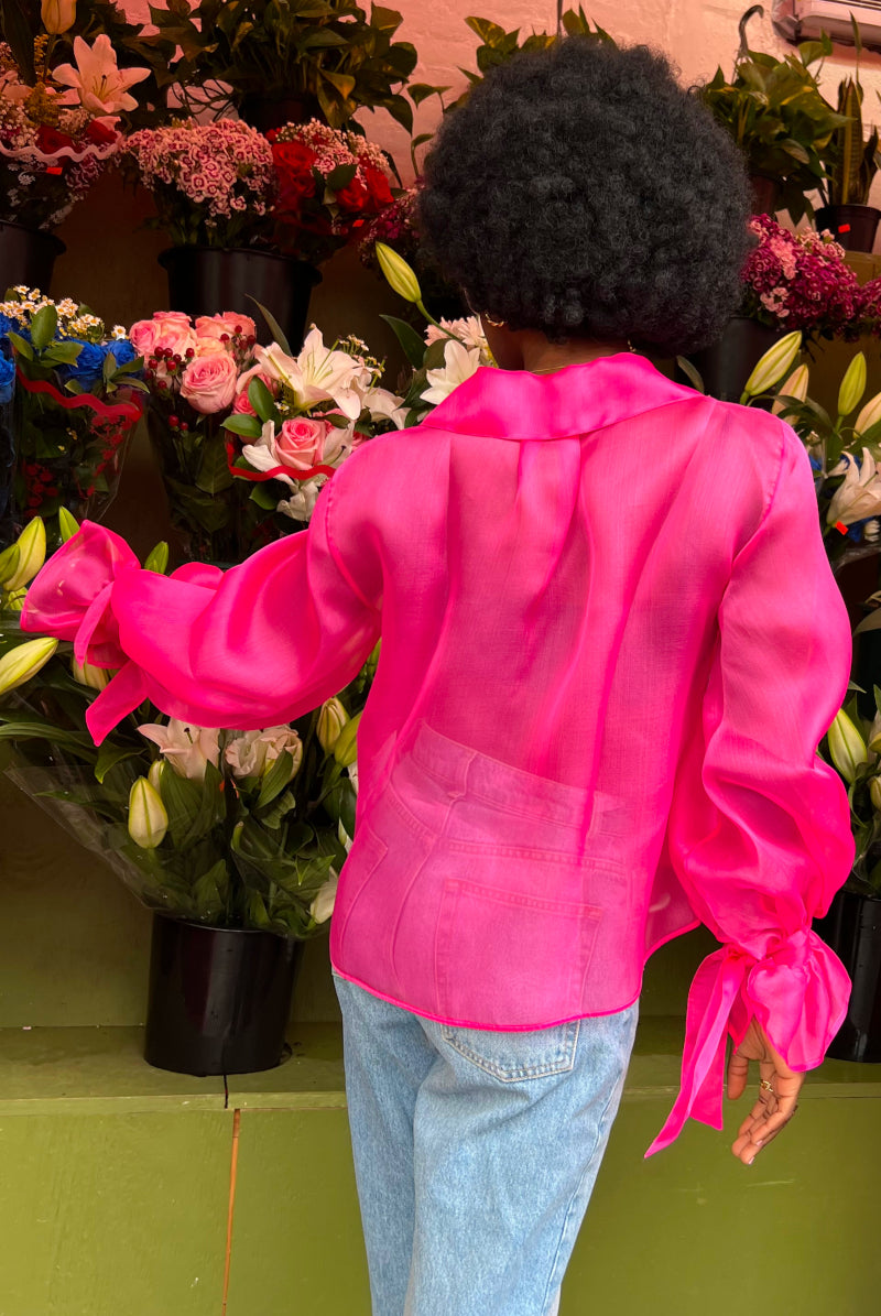 Rückenansicht einer afro-amerikanischen jungen Frau in einem Blumenladen, während sie eine blaue Jeans an hat und eine Seiden Organza Bluse in Hot Pink von Lola Tong.