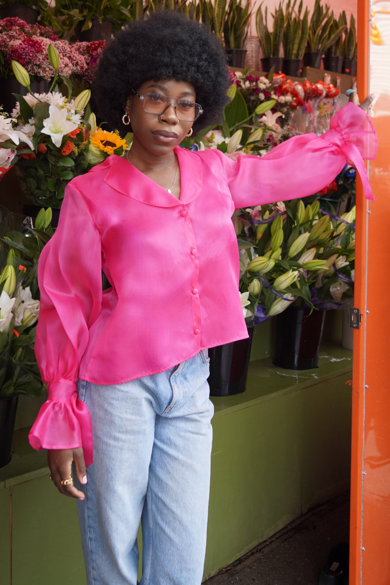 Eine junge modische afro-amerikanische Frau, die sich an einen Blumenladen anlehnt und gestylt ist in blauer Jeans und einer Seiden Organza Bluse in Hot Pink von Lola Tong. 