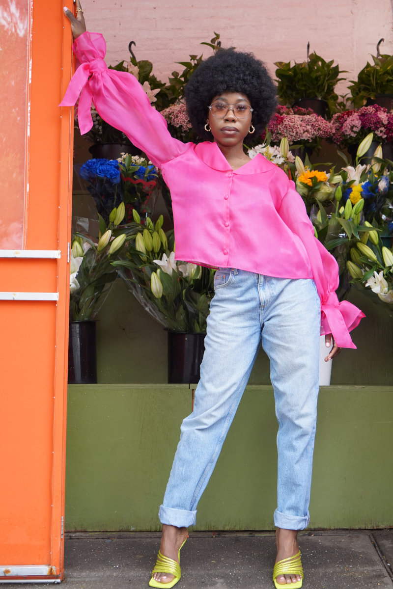 Eine junge modische afro-amerikanische Frau, die sich an einen Blumenladen anlehnt und gestylt ist in blauer Jeans und einer Seiden Organza Bluse in Hot Pink von Lola Tong und ihre Haare sind in einem wunderschönen Afro geschnitten.